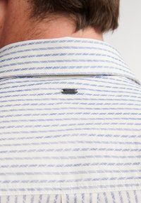 Camicia a righe con strisce orizzontali blu e bianche, caratterizzata da un colletto button-down e un piccolo etichetta nera cucita sul retro.