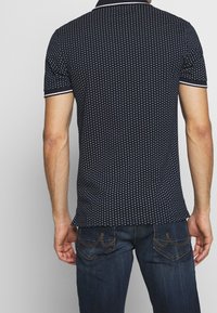 Navy polo shirt with white dot pattern, short sleeves, and contrasting collar and sleeve trim. Paired with dark denim jeans.