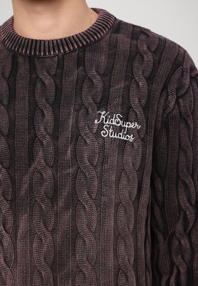 Pull en maille torsadée marron avec un col côtelé. Présente un logo brodé en blanc "KidSuper Studios" sur le côté gauche. Motif texturé sur l'ensemble.