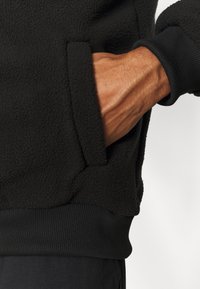 Main dans la poche d'une veste polaire noire avec poignets et taille côtelés, montrant un tissu texturé et un teint hâlé.