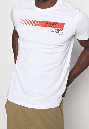 Homme portant un t-shirt blanc avec un texte graphique rouge et noir "CORE" et des chiffres sur la poitrine, associé à un pantalon beige.