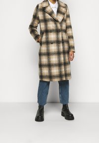 Cappotto scozzese doppiopetto in tonalità beige e marrone, realizzato in tessuto texturizzato. Caratterizzato da un colletto a revers e tasche laterali, abbinato a jeans blu e stivali neri.