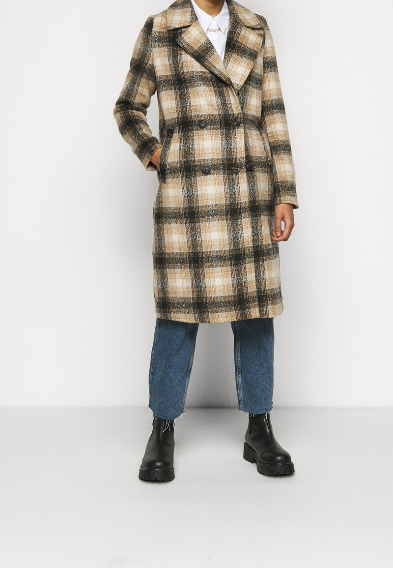 Cappotto scozzese doppiopetto in tonalità beige e marrone, realizzato in tessuto texturizzato. Caratterizzato da un colletto a revers e tasche laterali, abbinato a jeans blu e stivali neri.
