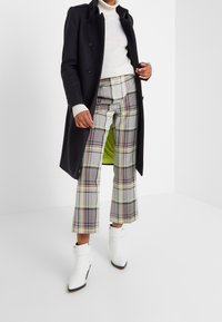 Abrigo largo negro, suéter de cuello alto blanco, pantalones de cuadros grises con toques de rosa y verde, y botines blancos con hebillas.