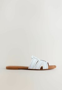 Sandale plate marron avec de larges brides en cuir blanc croisant le pied et un design à enfiler, ouvert à l’avant, sur un fond neutre.