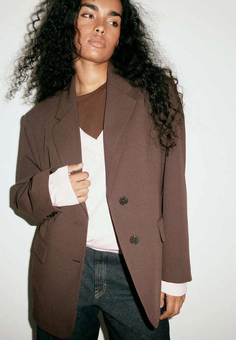 Femme aux longs cheveux bouclés portant un blazer marron oversize par-dessus des hauts blancs et marron superposés et un jean foncé, debout devant un fond uni.