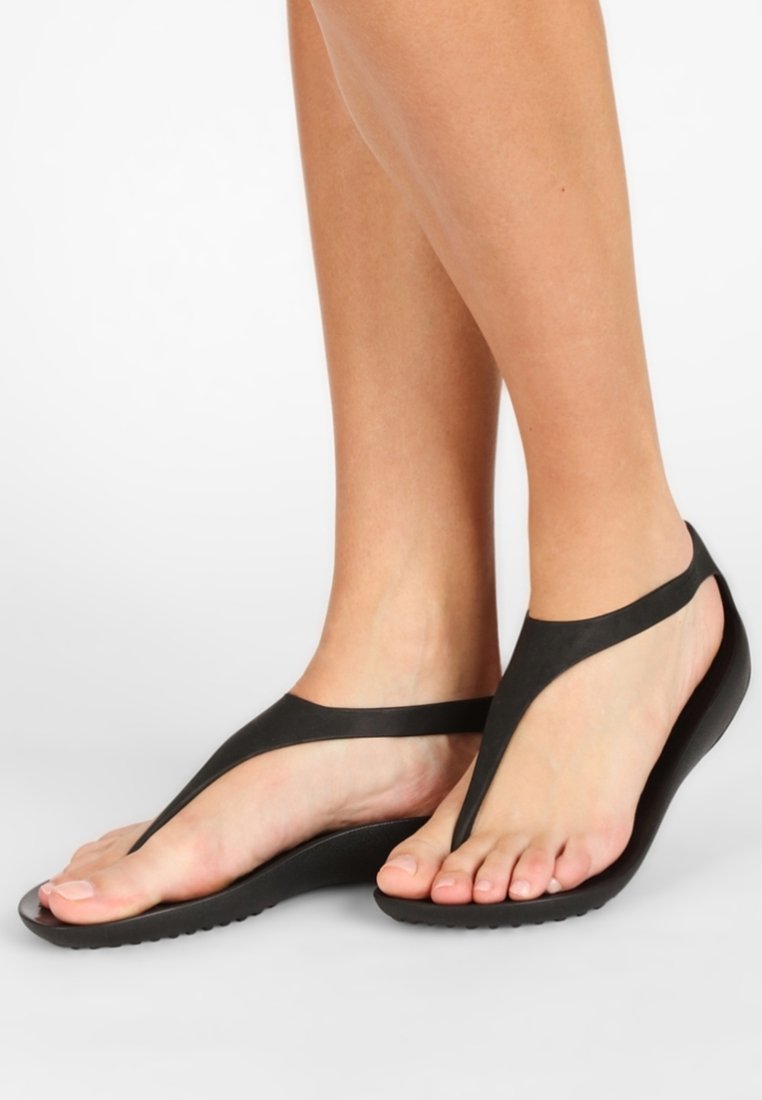 Pieds portant des tongs en caoutchouc noir avec une semelle courbée sur un fond blanc, montrant une vue latérale et supérieure de la chaussure.