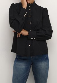 Blouse noire à manches longues avec un col montant, bordée de dentelle sur le devant, et boutons en perles. Le matériau semble doux et léger.