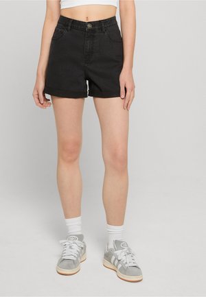 Inimene, kes kannab rullitud musta teksapükstega lühikesi pükse, valget lühikest toppi, valgeid sokke ja halli vabaajajalatsit, seisab ühevärvilise halli tausta ees.