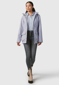 Giacca impermeabile viola chiaro con cappuccio, tasche frontali, indossata sopra una maglietta a righe e jeans scuri, completata da stivaletti beige alla caviglia.