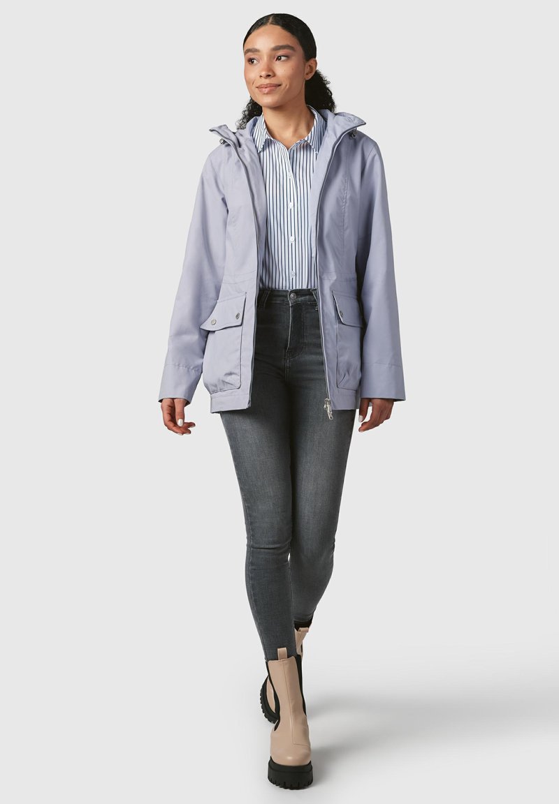 Giacca impermeabile viola chiaro con cappuccio, tasche frontali, indossata sopra una maglietta a righe e jeans scuri, completata da stivaletti beige alla caviglia.