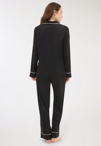 Donna in piedi a piedi nudi, rivolta di spalle, che indossa un top a maniche lunghe nero e pantaloni con rifiniture bianche sui polsini e sul colletto.