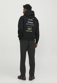 Sudadera negra con países impresos en varios colores en la parte posterior. Cuenta con un bolsillo tipo canguro y puños y dobladillo acanalados. Combinada con pantalones negros.