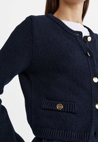 Navy knitted cardigan with a rounded neckline, gold buttons, and a front pocket. Features a textured fabric and ribbed hem.