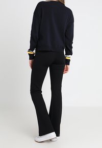 Granatowy sweter z dzianiny z żółtymi i białymi paskami na mankietach, w zestawieniu z czarnymi spodniami dzwonami i białymi sneakersami, widziane z tyłu.