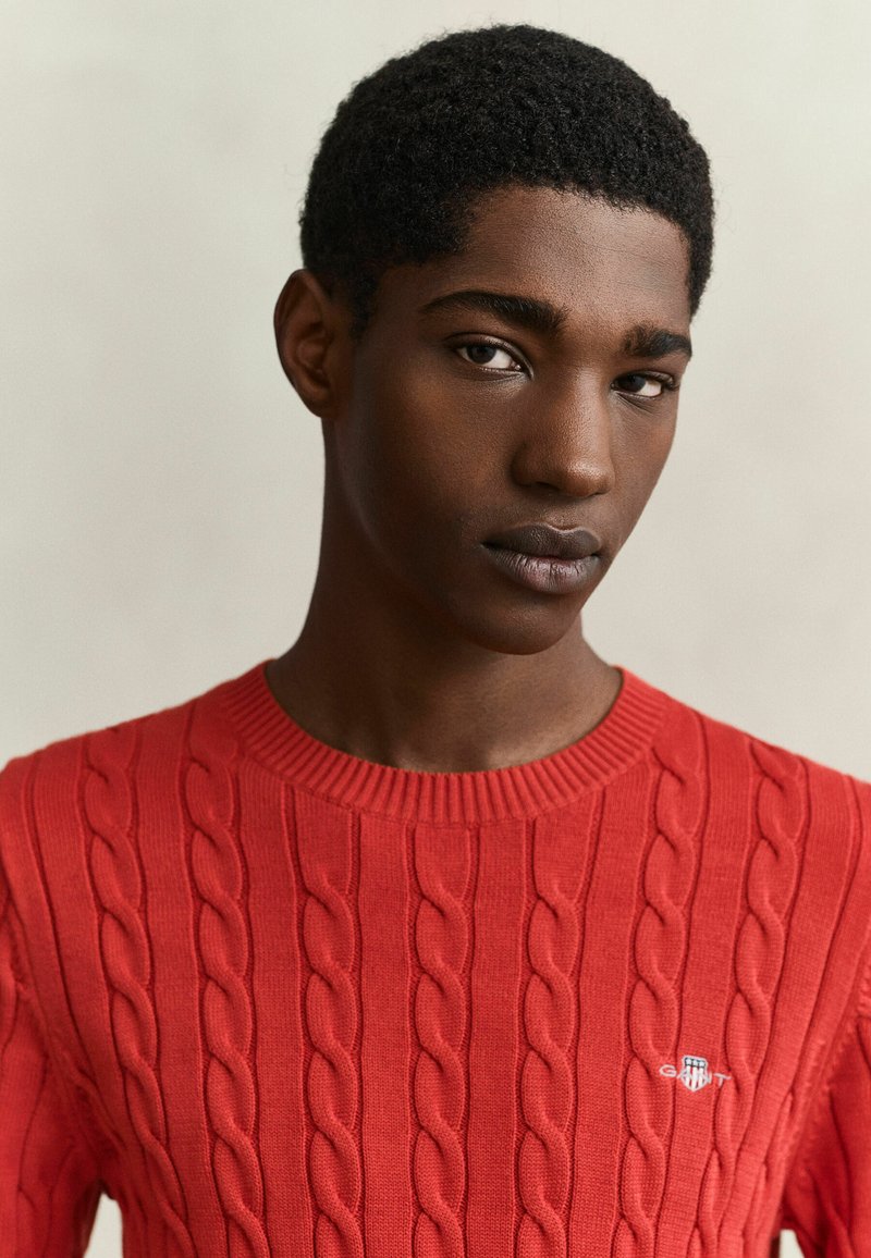 Young man with short curly hair wearing a red cable-knit sweater with a small logo on the chest, facing forward with a neutral expression.