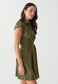 Abito verde oliva con maniche corte, colletto e vita accentuata da una cintura, caratterizzato da una morbida texture e un design a gonna plissettata.