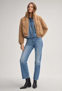 Brązowa kurtka z sztucznego futra o kroju cropped z teksturowanymi panelami, połączona z jasnoniebieską koszulą z dżinsu i prostymi dżinsami. Czarne botki do kostek dopełniają stylizację.