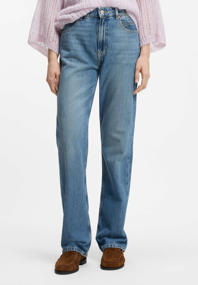 Jeans di denim azzurro chiaro con taglio a gamba dritta, caratterizzati da un design con cinque tasche e chiusura con bottone metallico, abbinati a mocassini in camoscio marrone.