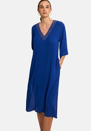 Femme portant une robe bleue ample jusqu'aux genoux avec des manches trois-quarts et un décolleté en V orné de perles, debout avec une main dans la poche.