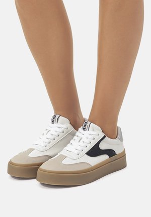 Zapatilla blanca y gris con acentos negros, materiales de cuero y gamuza, diseño de cordones plano y una suela de goma gruesa en color marrón claro.