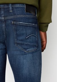 Modré džíny z denimu s klasickým designem pěti kapes, zadní kapsa má výrazný zakřivený steh a na pásku je kožená nášivka.