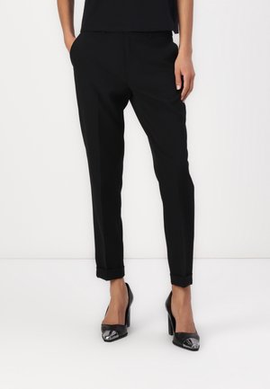 Vrouw met strakke zwarte cropped broek met omgeslagen pijpen en zwarte hoge hakken met metalen, puntige neuzen, staand met handen in de zakken.