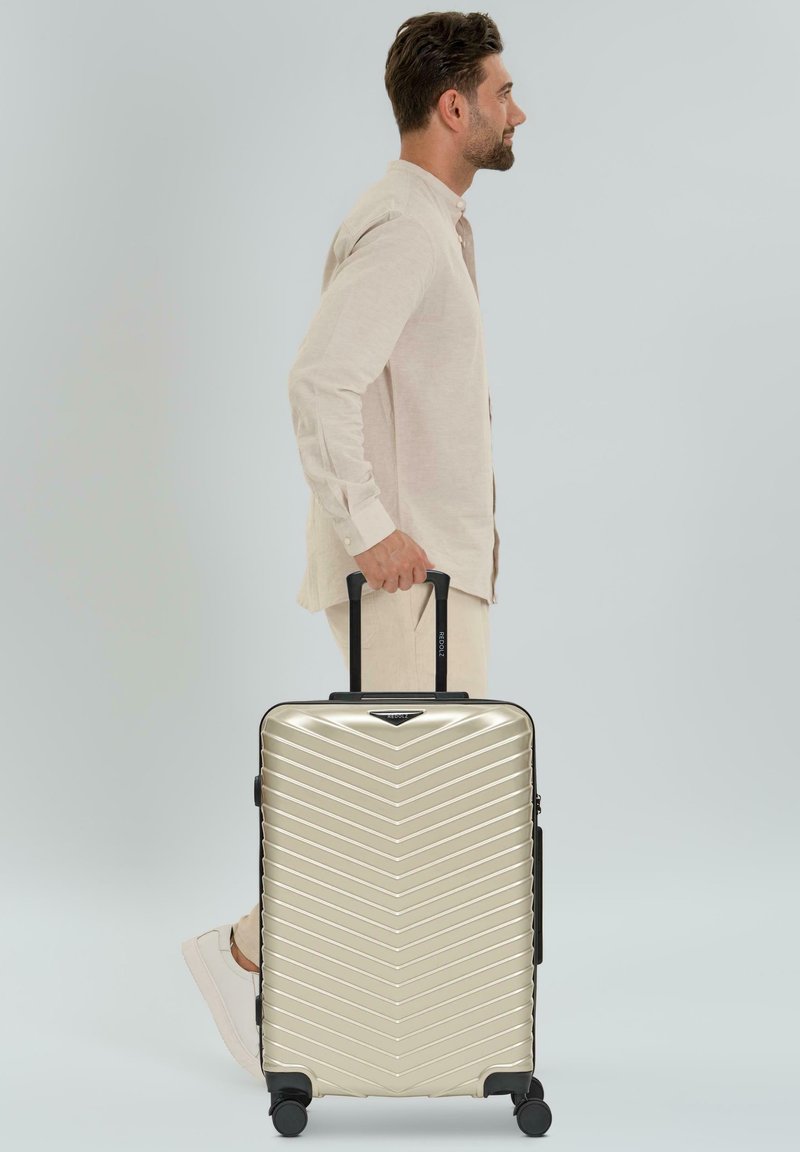 Man in beige outfit die zijwaarts loopt en de handgreep van een grote metalen gouden koffer met wielen vasthoudt tegen een vlakke achtergrond.