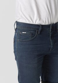 Sötétkék farmernadrág, amelyet betűrt fehér pólóval viselnek, elöl zsebekkel és övbújtatókkal.