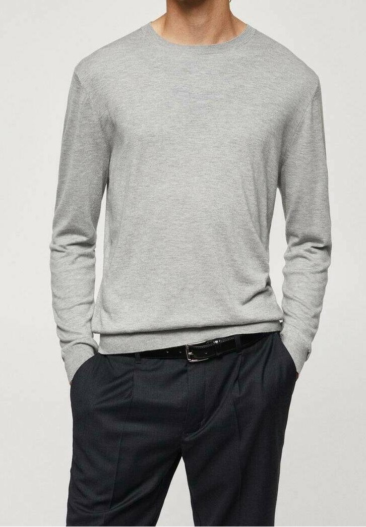Homme portant un pull léger à manches longues gris clair, légèrement rentré dans un pantalon habillé noir avec une ceinture noire, mains dans les poches.
