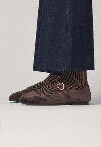 Scarpe in suede marrone con punta rotonda, dotate di una fascia e una fibbia rotonda dorata. Abbinate a calze corte a righe di colore scuro.