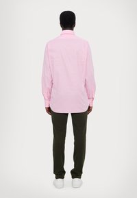 Man som vänder ryggen, iförd en långärmad ljusrosa rutiga skjorta, mörkgröna byxor och vita sneakers mot en enkel ljus bakgrund.