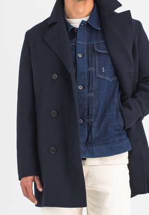 Un mannequin partiellement cadré porte un manteau croisé bleu marine par-dessus une veste en jean bleue, une chemise blanche et un pantalon crème.