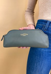 Persoon in blauwe jeans en een beige trui houdt een groene leren ritstas met textuur vast, waarop "Cresti Leder studio" in goud is geperst.