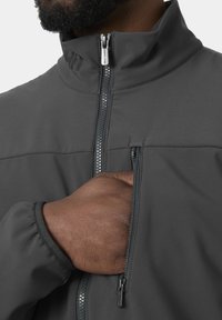 Persona con barba, que lleva una chaqueta oscura con la mano dentro del bolsillo en el pecho con cremallera, cuello parcialmente cerrado, sobre un fondo claro.