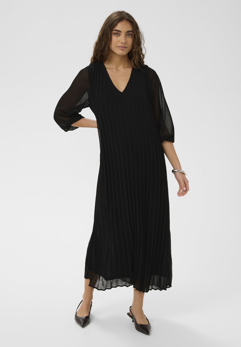 Femme portant une robe midi noire plissée à col en V avec des manches trois-quarts transparentes, ainsi que des talons slingback noirs à bout pointu, debout sur un fond blanc.