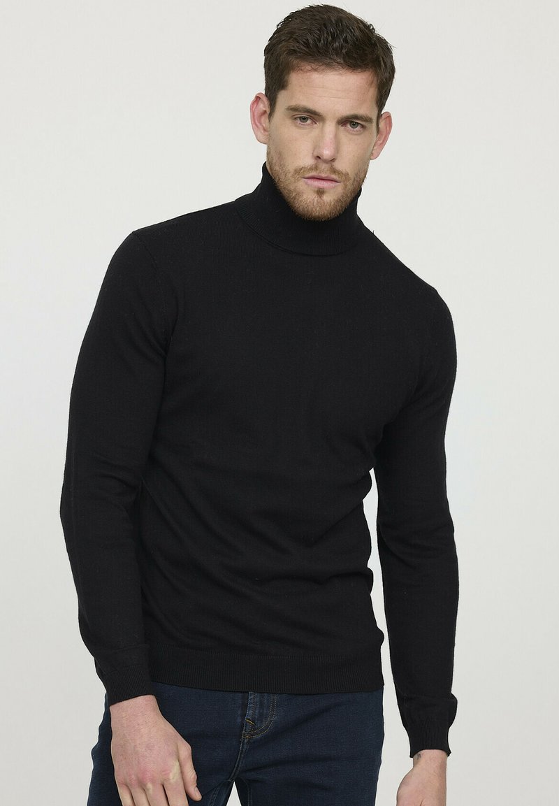 Pull à col roulé noir en tissu côtelé, au design ajusté, manches longues et col haut et serré. Porté avec un jean bleu foncé.