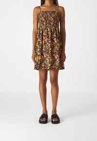 Frau trägt ein ärmelloses, knielanges Blumenkleid mit gerafftem Oberteil und schwarzen Sandalen vor einem schlichten weißen Hintergrund.