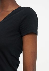 Κοντινό πλάνο του πάνω μέρους του κορμού και του βραχίονα ενός ατόμου που φοράει εφαρμοστή μαύρη κοντομάνικη μπλούζα με V λαιμόκοψη, σε απλό λευκό φόντο.