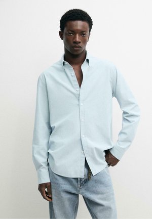 Jeune homme portant une chemise bleu pâle à boutons et un jean bleu clair, debout devant un fond blanc uni avec une main sur la hanche.