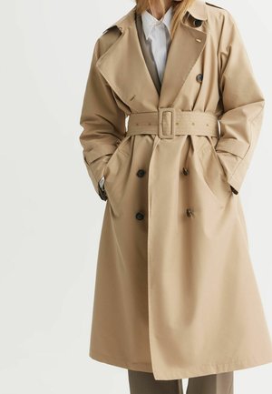 Kobieta nosząca beżowy trencz z paskiem, z dwurzędowymi guzikami, kieszeniami i białą koszulą z kołnierzykiem pod spodem, stojąca z rękami w kieszeniach.