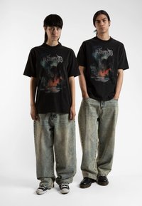 Črne grafične majice s kratkimi rokavi vsebujejo velik, barvit dizajn z zmajem. Nosijo se z ohlapnimi, svetlo modrimi, postaranymi baggy jeans hlačami in kontrastnimi supergami.