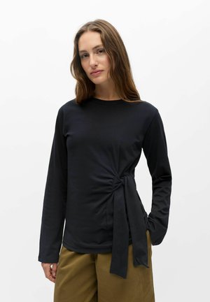 Femme aux longs cheveux bruns portant un haut noir à manches longues avec un lien sur le côté et un pantalon marron clair sur fond blanc uni.