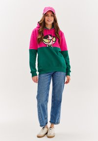 Hoodie v růžové a zelené barvě s grafikou karikatury, s kapucí na šňůrky. V kombinaci s světle modrými džíny a bílými teniskami.