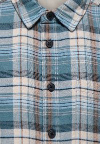 Chemise à carreaux bleu, beige et marron avec des boutons noirs et une poche poitrine, montrée en gros plan du col à la taille.