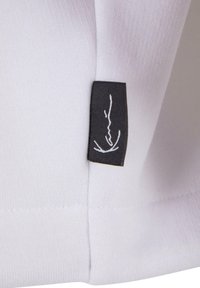 Biely bavlnený látka s čiernym štítkom, ktorý zobrazuje minimalistický biely lineárny kresbu, vyšitý na šve. Textúra vyzerá hladko a jemne.