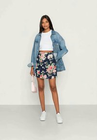 Mulher a usar um top cropped branco, saia floral azul-marinho, um casaco de ganga oversized, sapatilhas brancas, a segurar uma pequena mala rosa, de pé contra um fundo simples.