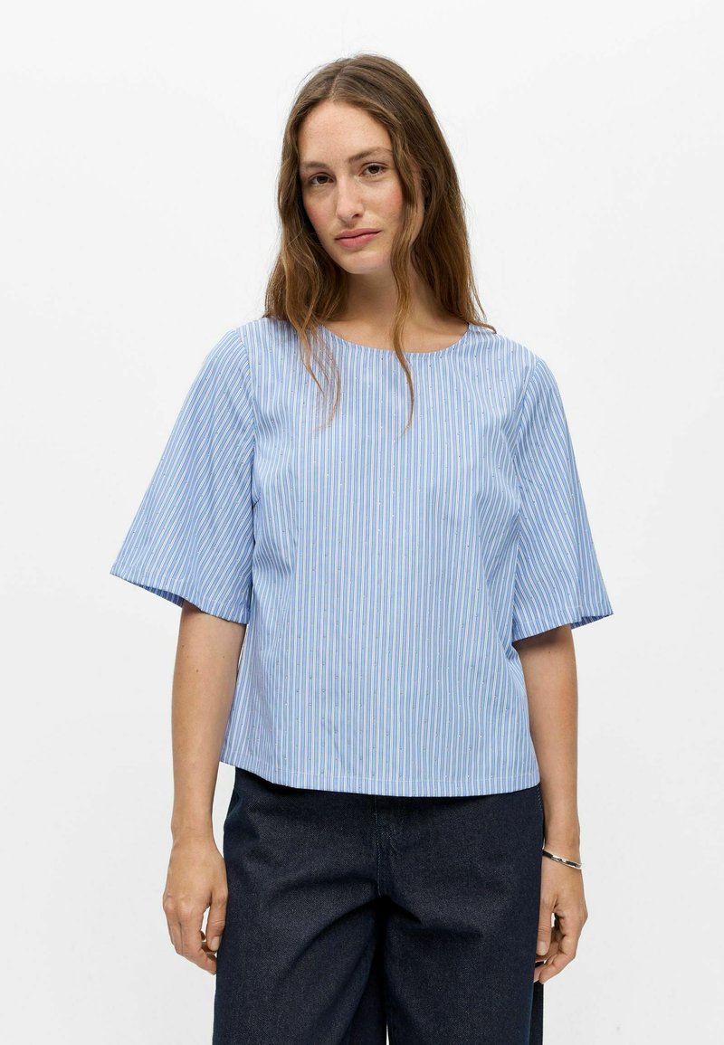 Jeune femme aux longs cheveux portant un haut à manches courtes rayé verticalement bleu et blanc et un pantalon foncé sur un fond uni.