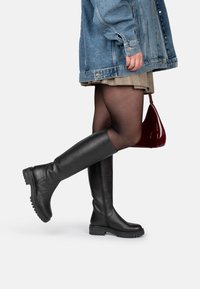Botas de cuero negras hasta la rodilla con un acabado texturizado, suela gruesa y estriada, y un brillo sutil, combinadas con una chaqueta de mezclilla y una falda de cuadros.