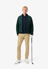 Veste rembourrée verte avec fermeture éclair, sous un vêtement bleu marine, pantalon beige, chaussures blanches et un club de golf. Comprend un logo sur la poitrine et la capuche.
